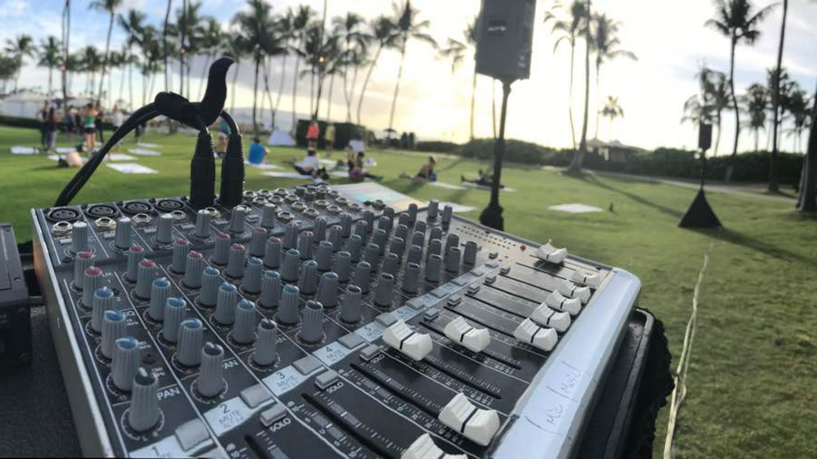 Allen and Heath QU16 digital mixing console with illuminated green faders and blue touchscreen at an outdoor evening event on grass with palm trees and relaxed participants in the background